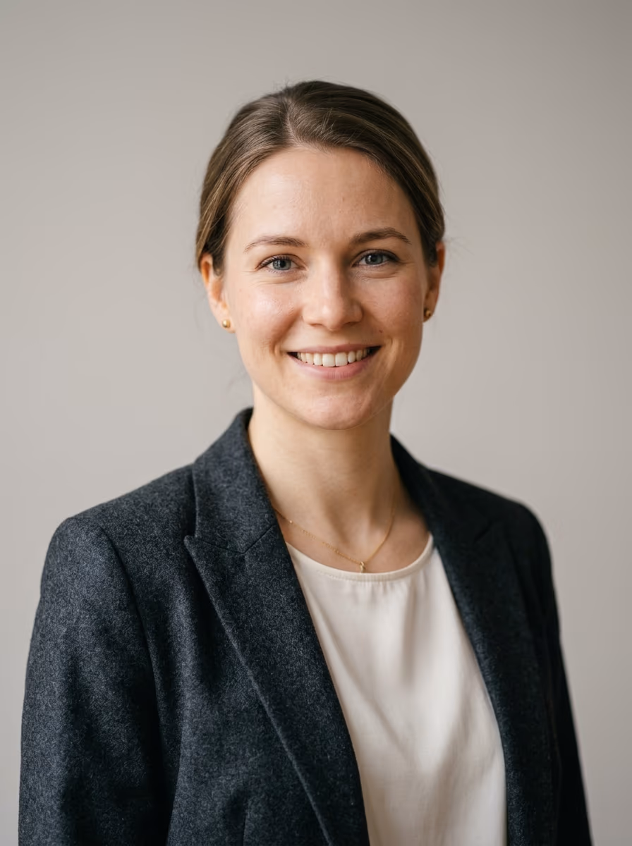 Portrait of a smiling woman in a charcoal blazer and cream top against a plain light wall