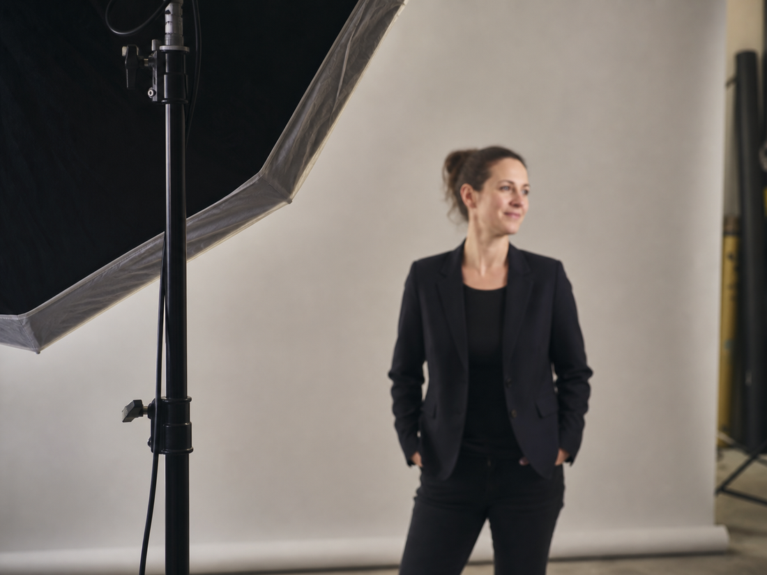 Blick von der Seite in ein Fotostudio, links im Bild eine Softbox auf einem Stativ,
            dahinter unscharf eine Frau im schwarzen Blazer vor hellgrauem Hintergrund