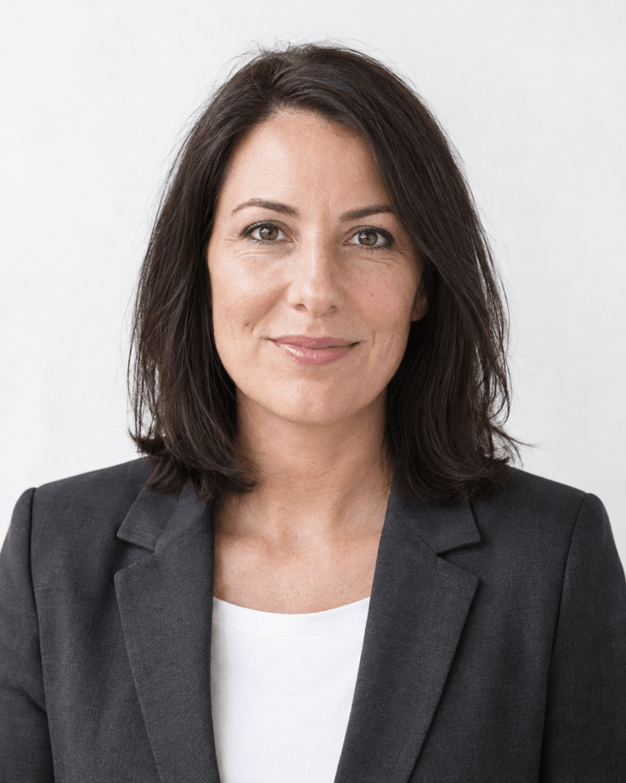 Bewerbungsfoto einer Frau um vierzig mit schulterlangen dunklen Haaren in dunkelgrauem Blazer über weißem Top vor cremeweißem Hintergrund, Blick in die Kamera