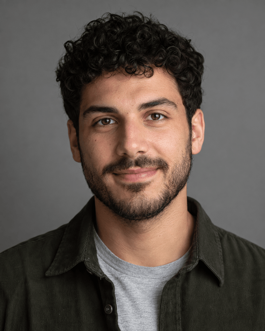 Bewerbungsfoto eines jungen Mannes mit kurzen schwarzen Locken und kurzem Bart in dunkelgrünem Overshirt über grauem T-Shirt vor mittelgrauem Hintergrund, leichtes Lächeln