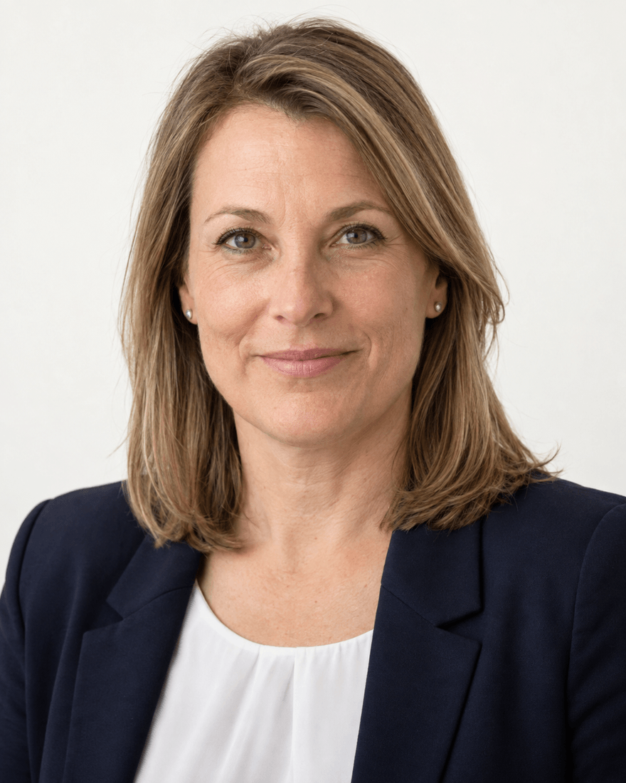 Frau Mitte vierzig mit schulterlangem dunkelblondem Haar im dunkelblauen Blazer über weißer Bluse vor cremeweißem Hintergrund, freundlicher fester Blick