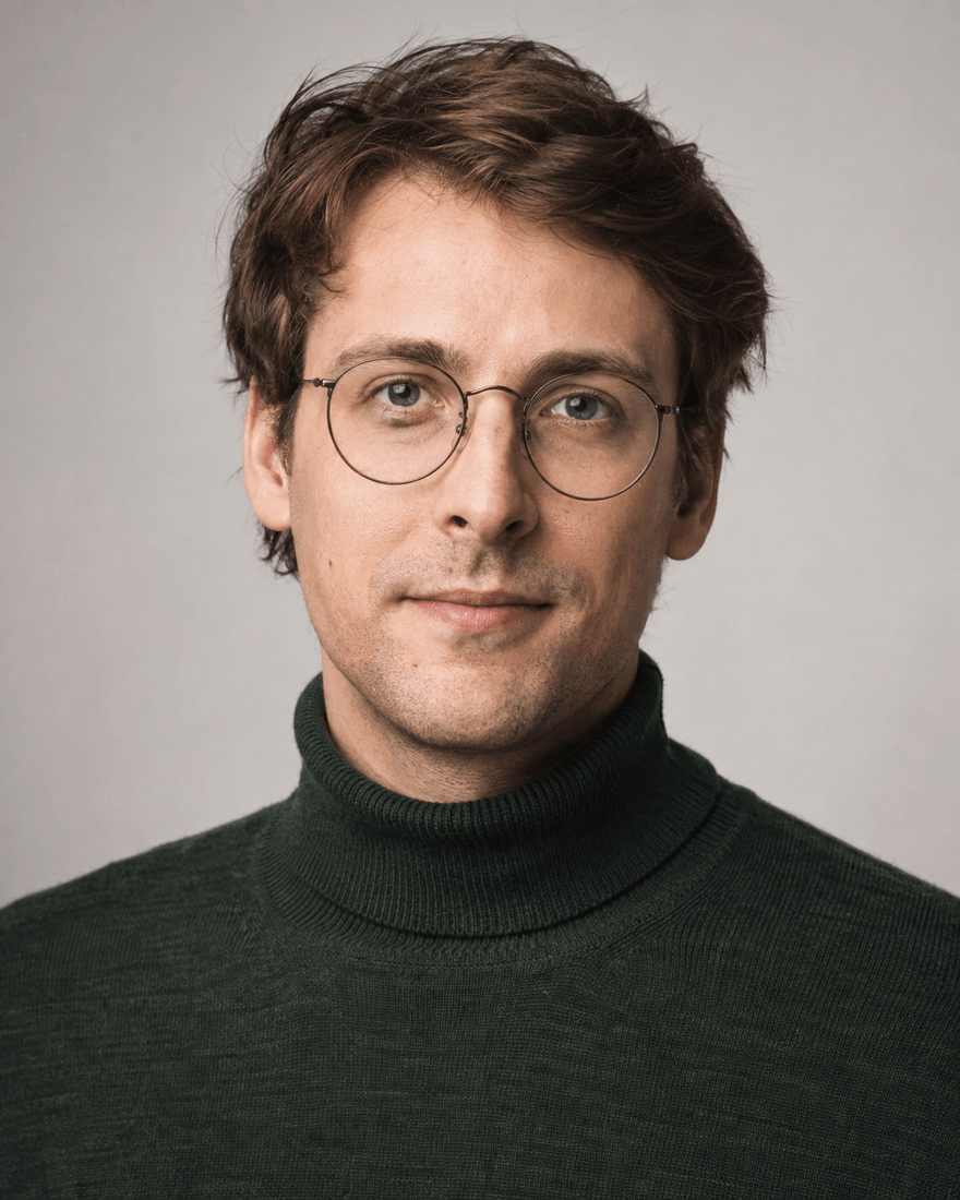 Mann Ende zwanzig mit halblangem braunem Haar und runder Brille im dunkelgrünen Rollkragenpullover vor warm grauem Hintergrund, aufmerksamer Blick