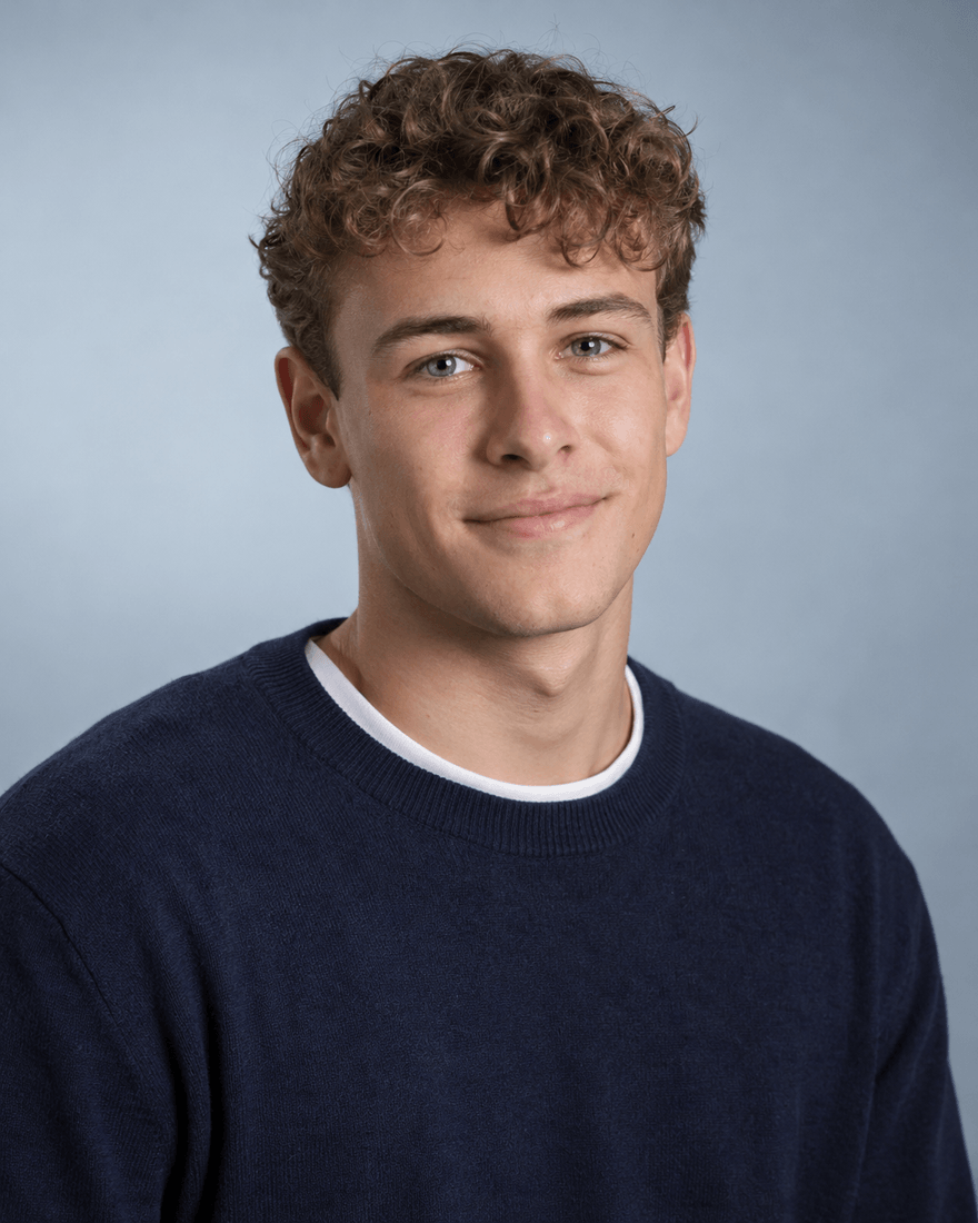 Junger Mann um die neunzehn mit kurzem lockigem dunkelblondem Haar in marineblauem Pullover vor einer hellen blaugrauen Studiowand, entspannter neugieriger Ausdruck