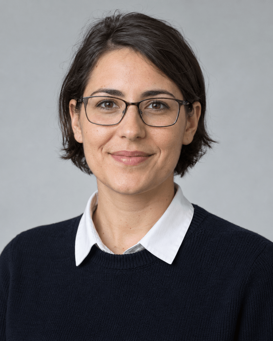 Bewerbungsfoto einer Frau Anfang dreißig mit kurzem dunkelbraunem Haar und schmaler dunkler Brille in dunkelblauem Pullover mit weißem Kragen vor hellgrauem Hintergrund, freundlicher Blick