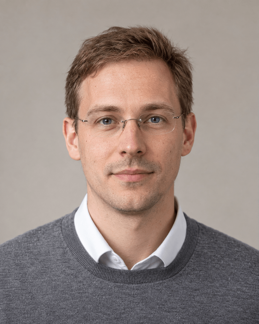 Mann um dreißig mit kurzem hellbraunem Haar und randloser Brille im mittelgrauen Pullover über weißem Hemdkragen vor hellem warmgrauem Studiohintergrund, ruhiger Blick in die Kamera