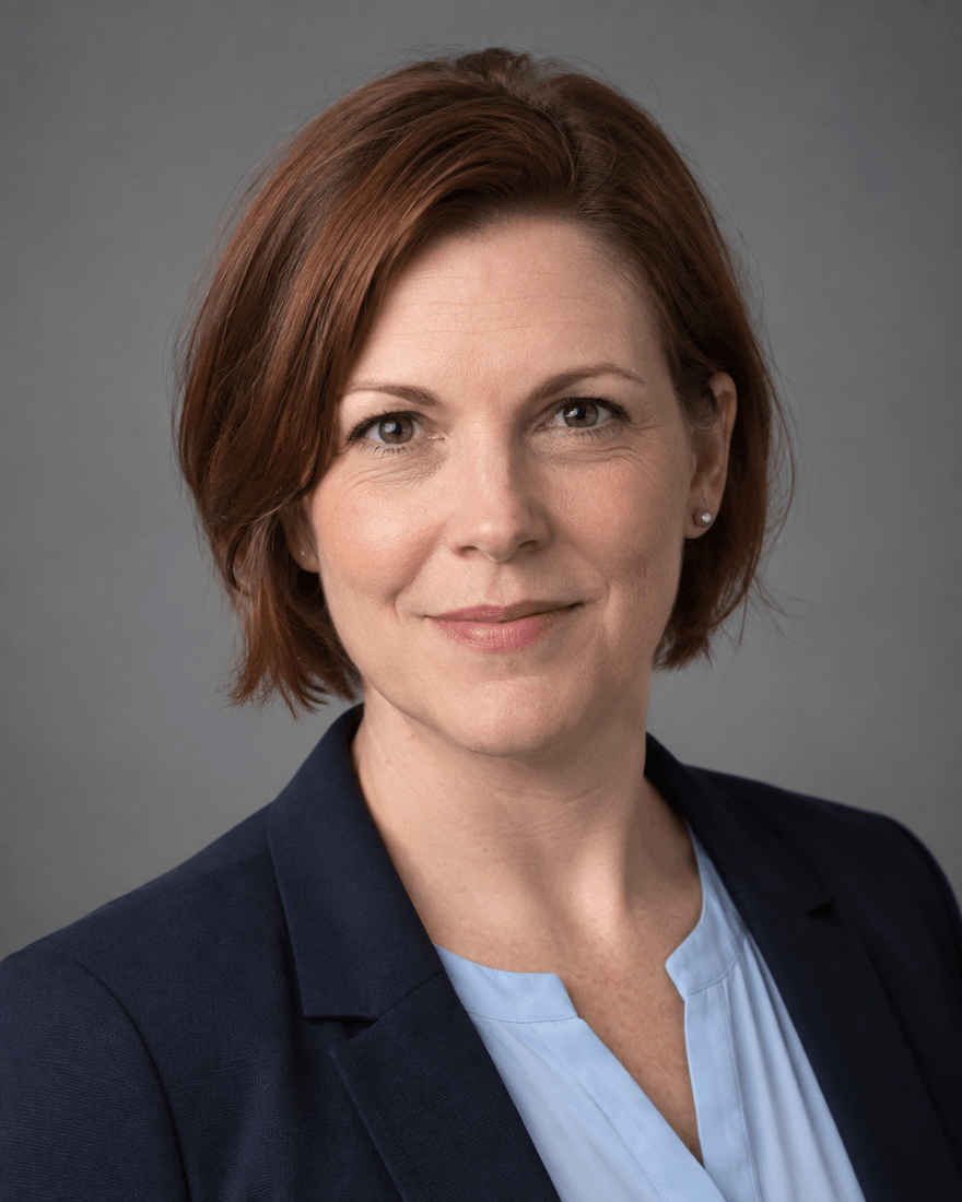 Frau um vierzig mit kinnlangem rotbraunem Haar im dunkelblauen Blazer über hellblauer Bluse vor mittelgrauem Studiohintergrund, freundlicher, sicherer Blick