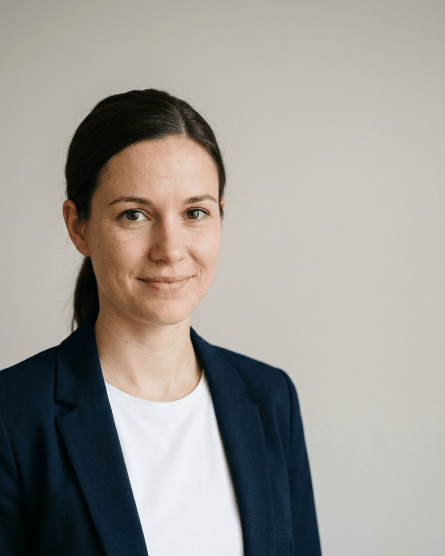 Frau Ende zwanzig mit dunklem Haar im niedrigen Zopf im dunkelblauen Blazer über weißem Rundhalsshirt, links im Bild, rechts ein breiter leerer Streifen heller Wand für Text