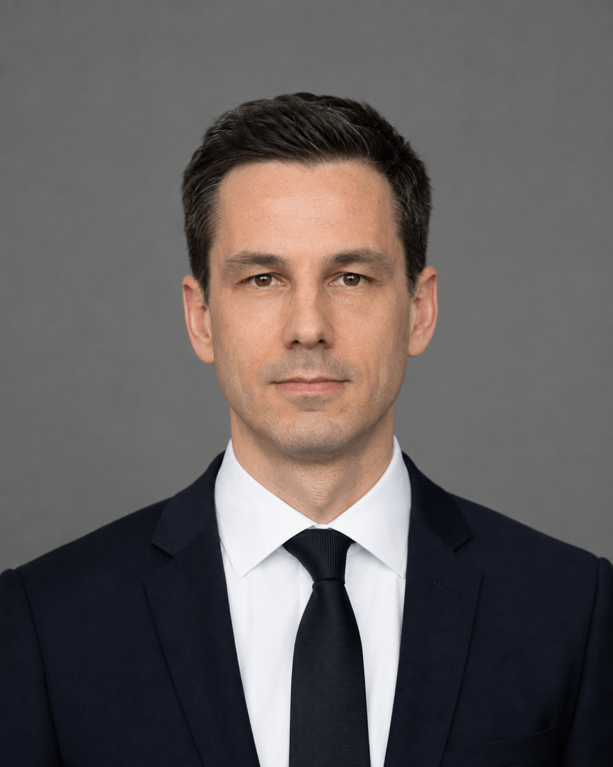Mann Mitte dreißig im dunkelblauen Anzug mit weißem Hemd und schmaler Krawatte vor einem gleichmäßig grauen Hintergrund, Kopf und Schultern mittig im Bild