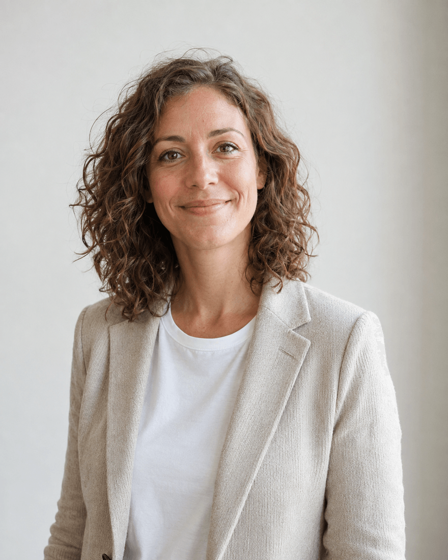 Frau Anfang dreißig mit lockigem braunem Haar im hellbeigen Strickblazer über weißem Shirt vor einer hellen cremefarbenen Wand, lächelnd