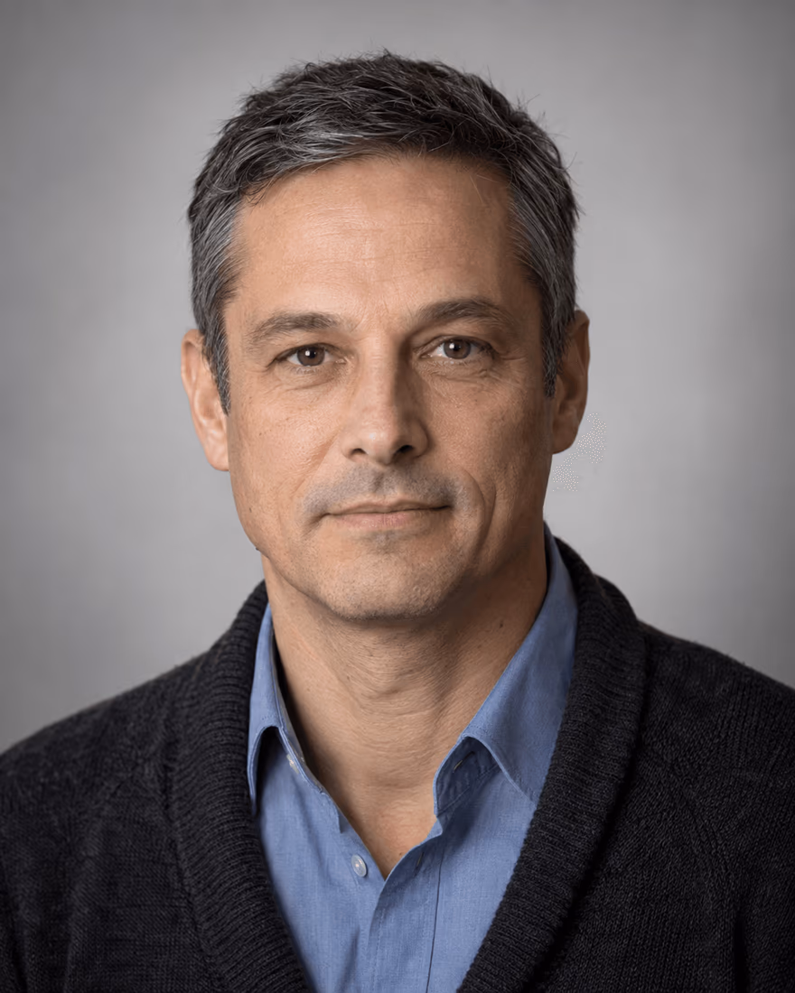 Retrato de un hombre de cabello canoso con jersey oscuro y camisa azul sobre un fondo gris claro