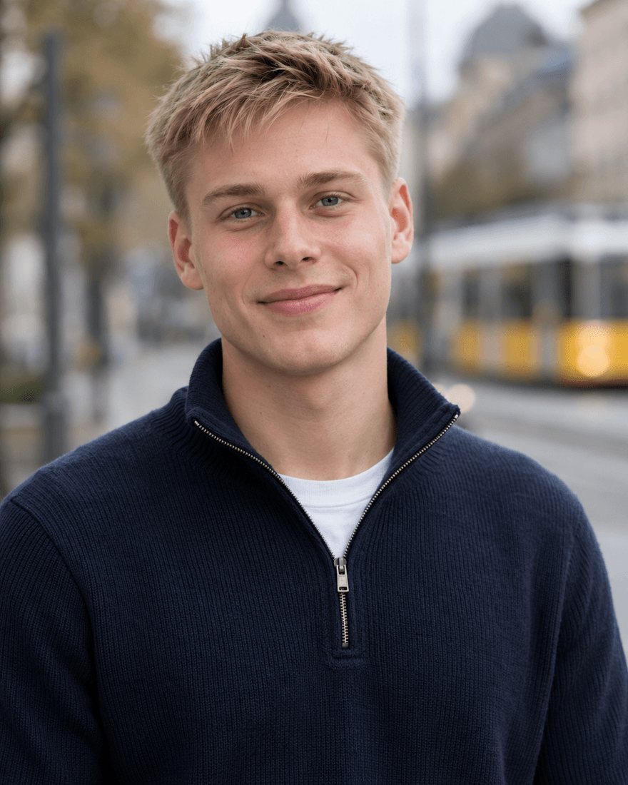 Bewerbungsfoto eines jungen Mannes von etwa zwanzig Jahren mit kurzem glattem blondem Haar in dunkelblauem Pullover mit kurzem Reißverschluss vor unscharfer Straße mit einer gelb-weißen Straßenbahn im Hintergrund