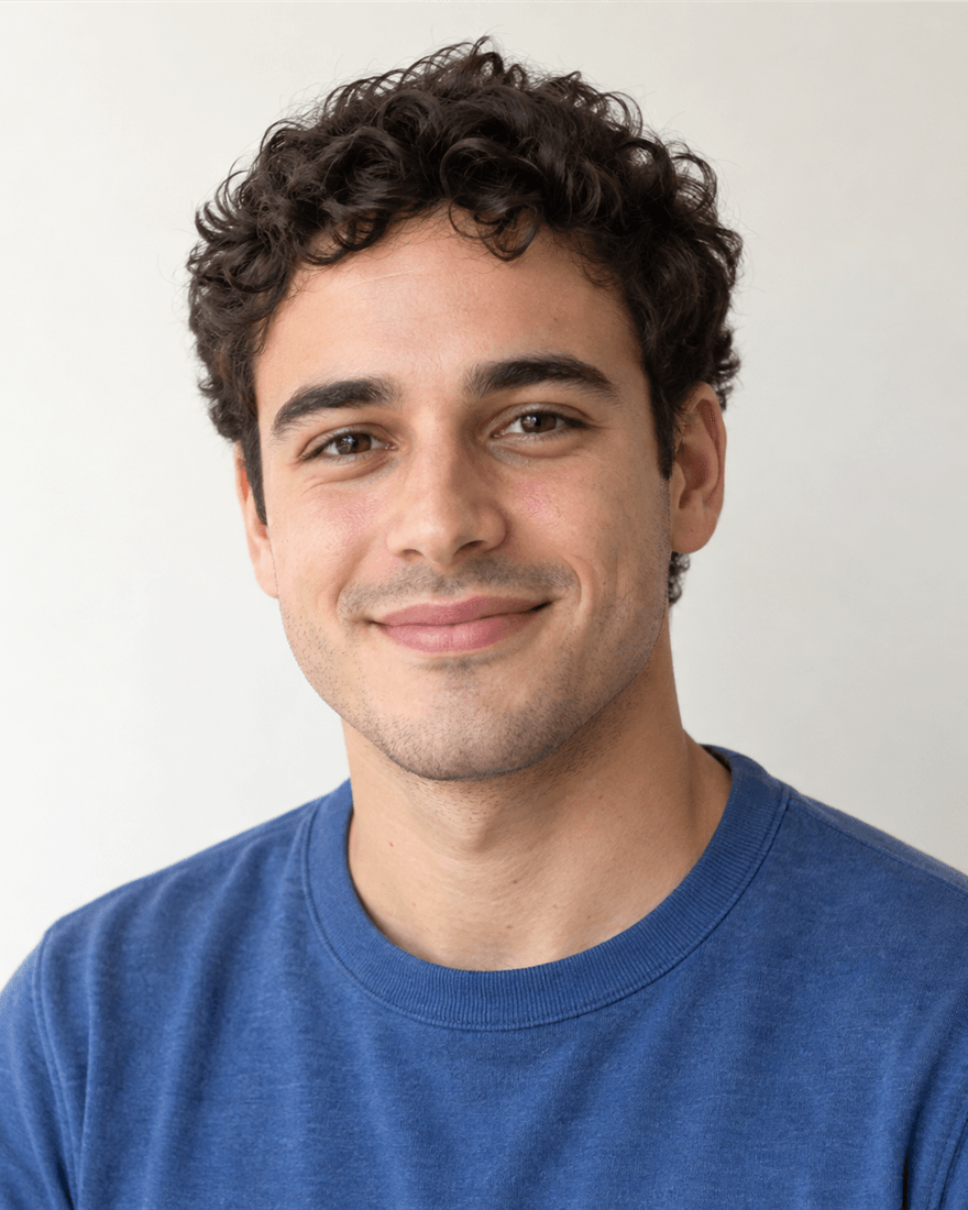 Junger Mann um 24 mit kurzem dunklem Lockenhaar im schlichten mittelblauen Shirt vor cremeweißem Hintergrund, Bewerbungsfoto für die Pflegeausbildung an der Universitätsmedizin Mainz