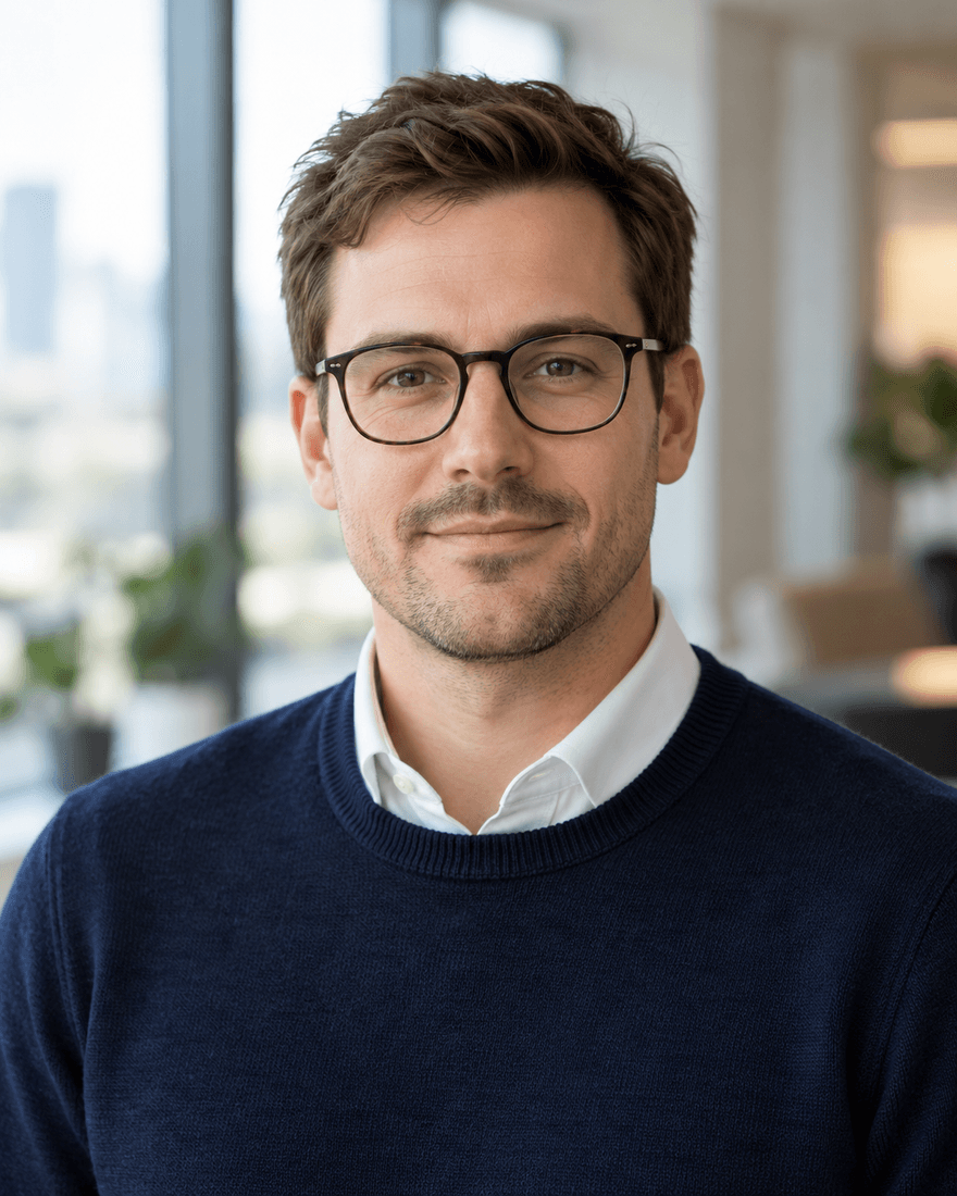 Bewerbungsfoto eines Mannes von etwa vierunddreißig Jahren mit kurzen braunen Haaren und Brille in dunkelblauem Pullover über weißem Hemdkragen vor einem unscharfen modernen Büro mit Fenster