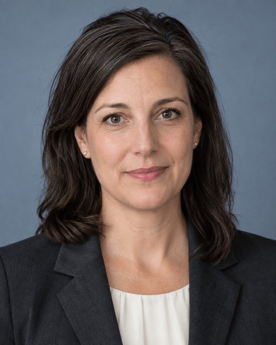 Bewerbungsfoto einer Frau Mitte vierzig mit schulterlangem dunklem Haar im anthrazitfarbenen Blazer über cremefarbener Bluse vor blaugrauem Hintergrund