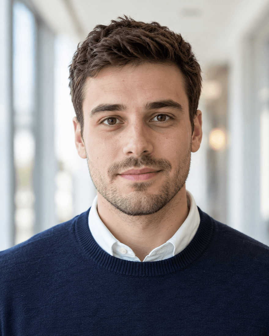 Mann um die sechsundzwanzig mit kurzen dunkelbraunen Haaren und gepflegtem Dreitagebart, weißes Hemd unter marineblauem Pullover, vor einem hellen unscharfen Flur