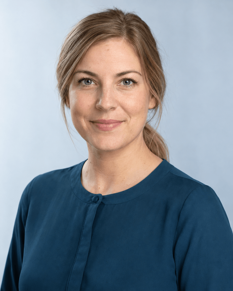 Frau um die vierunddreißig mit dunkelblondem Zopf und petrolblauer Bluse vor hellblaugrauem Hintergrund, entschlossener freundlicher Blick