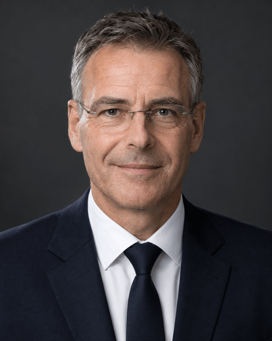 Bewerbungsfoto eines Mannes um fünfzig mit grau meliertem Haar und randloser Brille in dunkelblauem Anzug mit Krawatte vor dunkelgrauem Hintergrund