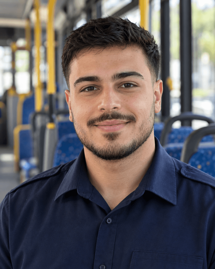 Bewerbungsfoto eines jungen Mannes von etwa einundzwanzig Jahren mit kurzen schwarzen Haaren und leichtem Bart in dunkelblauem Uniformhemd ohne Logo vor dem unscharfen Innenraum eines Stadtbusses mit blauen Sitzen, offen und freundlich