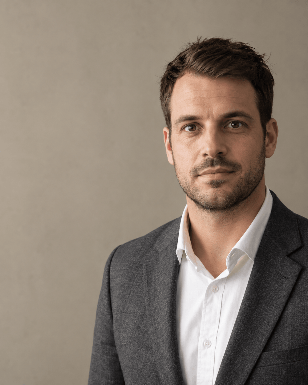 Mann Mitte dreißig mit kurzem dunkelbraunem Haar und kurzem Bart im dunkelgrauen Blazer über weißem Hemd, ruhiger Blick in die Kamera vor sandsteinfarbener Studiowand