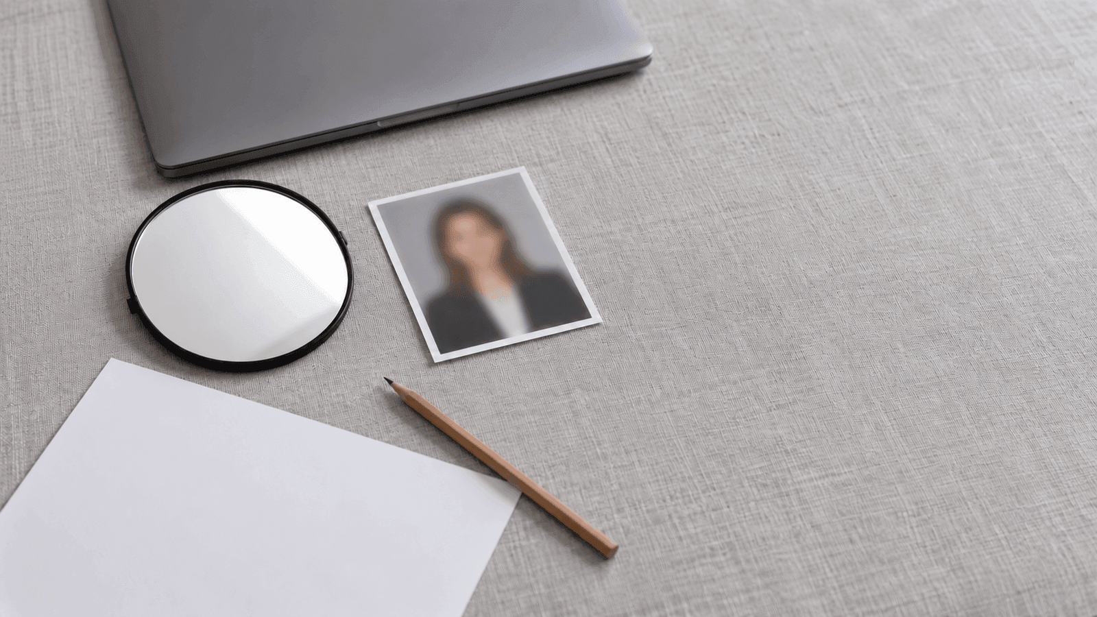 Auf einem hellgrauen Tisch liegen ein kleiner runder Handspiegel, ein unscharfer Porträtabzug, ein leeres weißes Blatt, ein Bleistift und ein zugeklappter Laptop