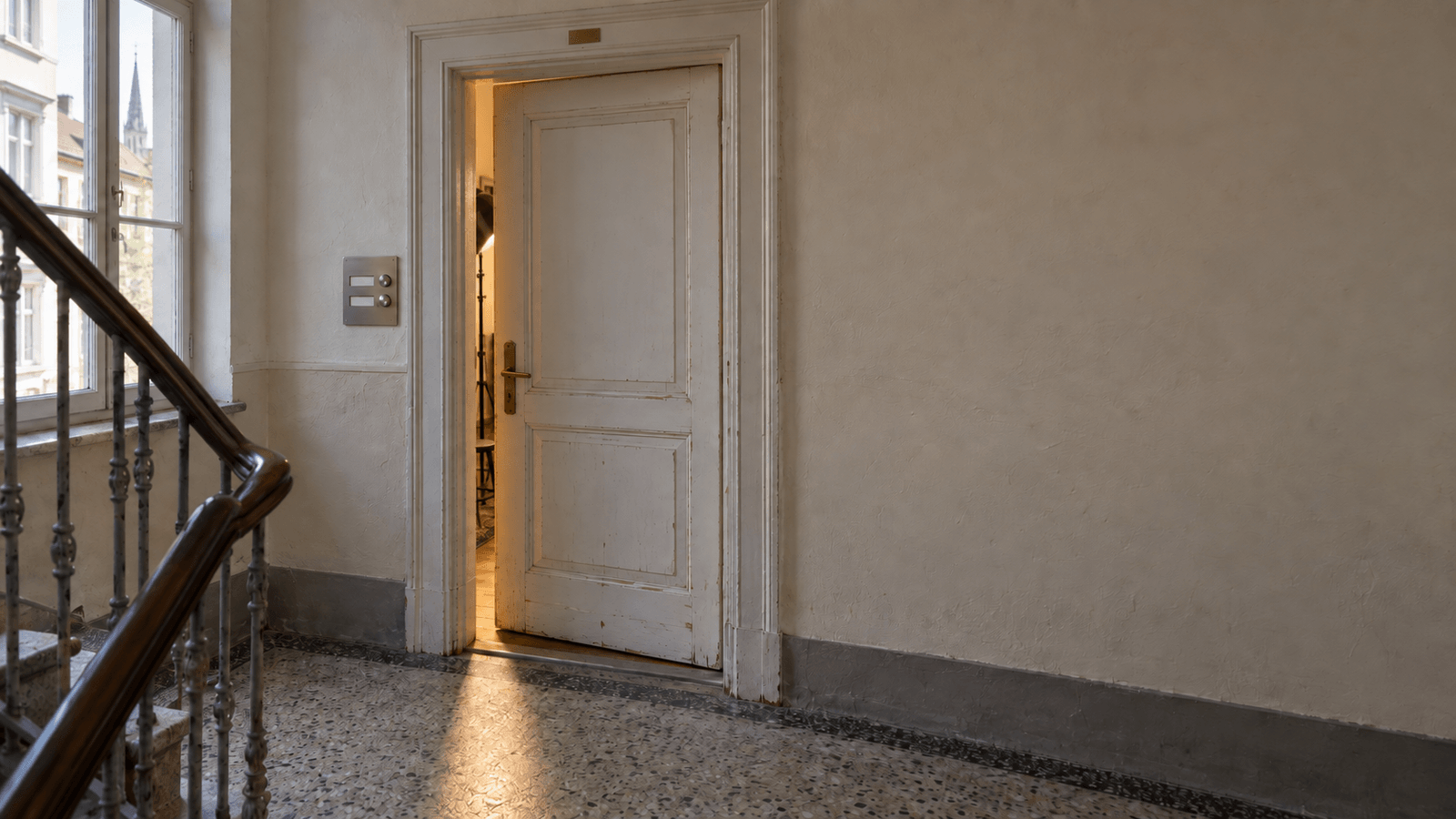 Weiße Altbautür eines Fotostudios steht einen Spalt offen, warmes Licht fällt auf Terrazzoboden, links an der Wand ein Klingelschild aus Edelstahl mit zwei leeren Namensschildern