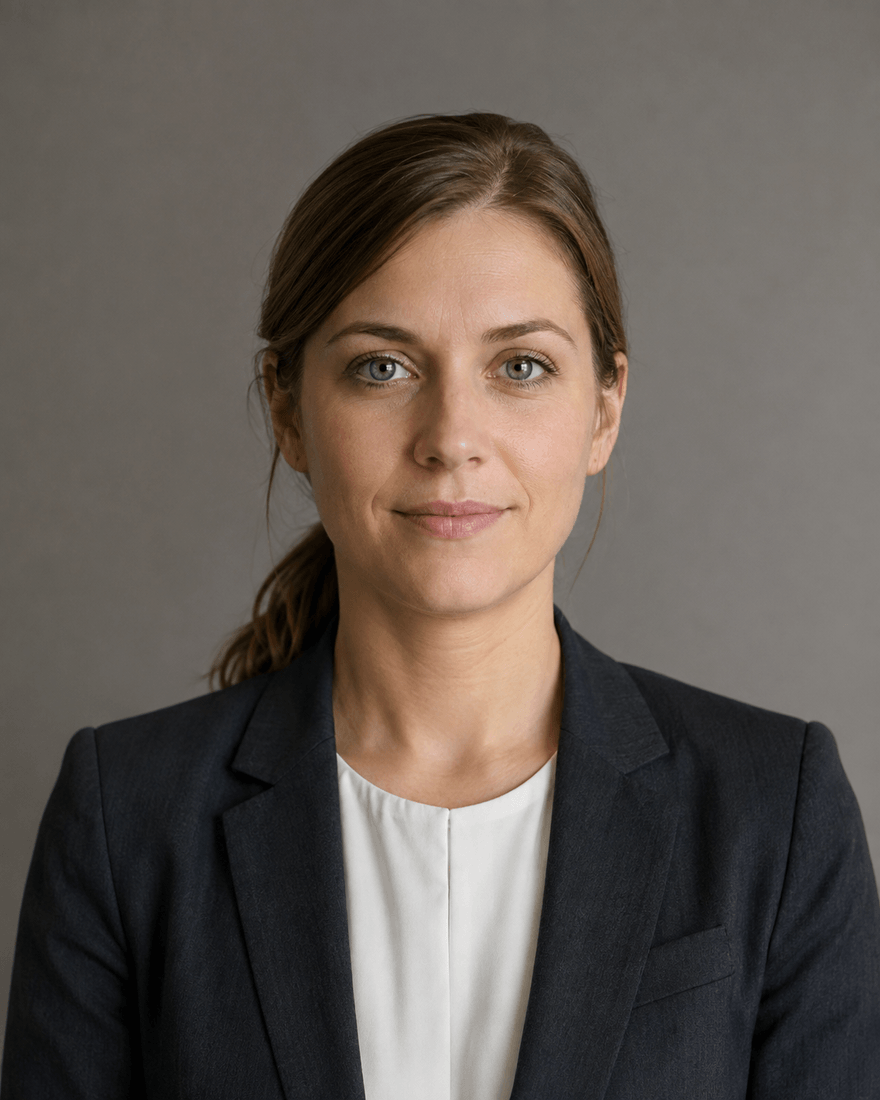 Junge Frau im dunklen Blazer über weißer Bluse vor einer warmgrauen Studiowand, ruhiger Blick in die Kamera