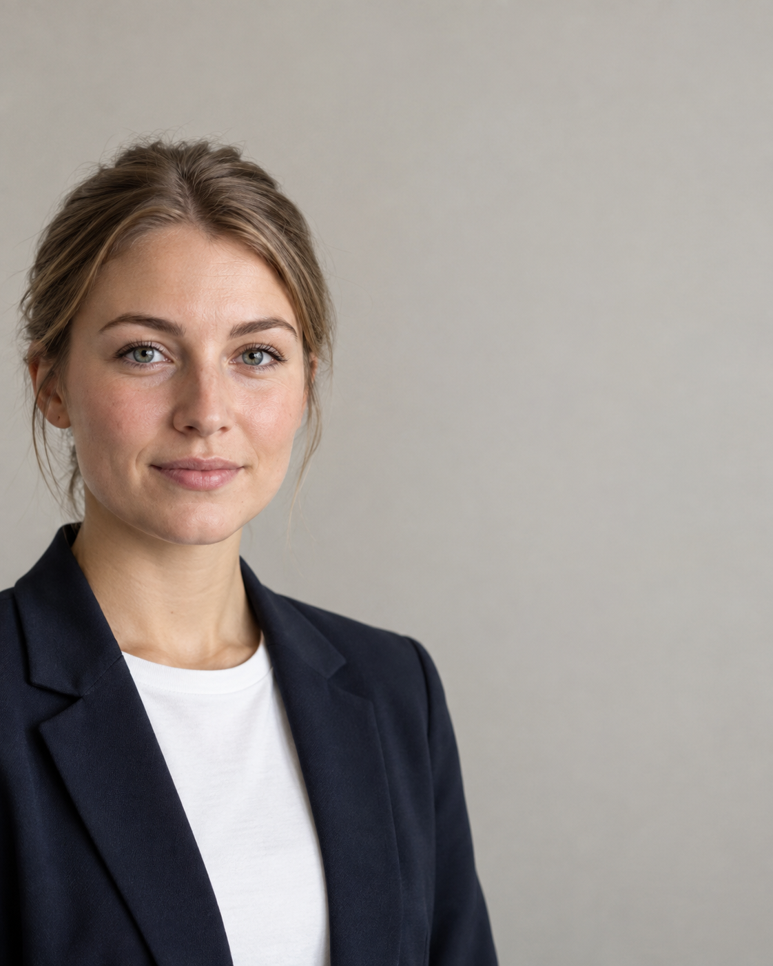 Junge Frau Anfang zwanzig mit zurückgebundenem dunkelblondem Haar im dunkelblauen Blazer über weißem T-Shirt vor hellgrauer Studiowand, ruhiger Blick in die Kamera