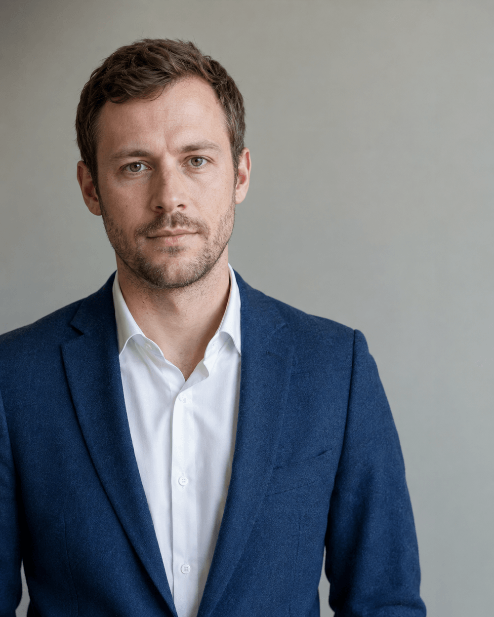 Mann Anfang dreißig im blauen Blazer über weißem Hemd vor einer hellgrauen Studiowand, ruhiger Blick in die Kamera