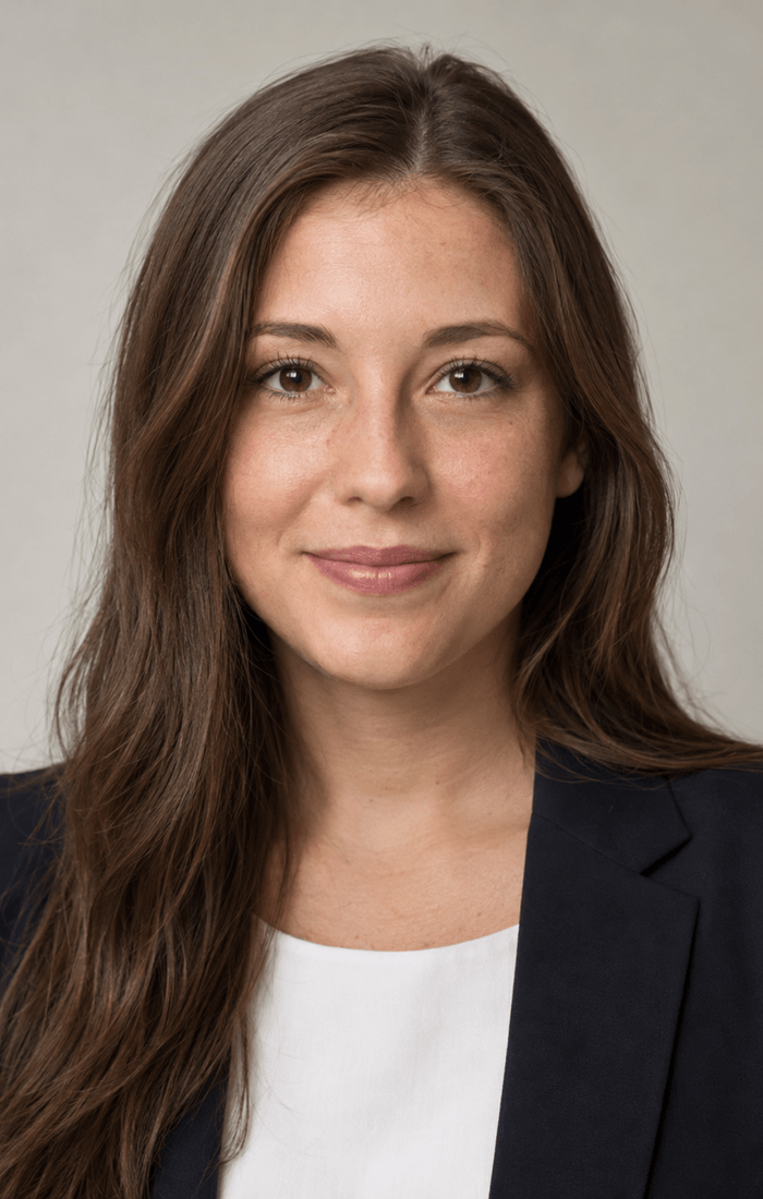 Bewerbungsfoto einer Frau Mitte zwanzig mit braunem Haar im dunkelblauen Blazer über weißer Bluse vor hellgrauem Hintergrund, im Seitenverhältnis 7 zu 11
