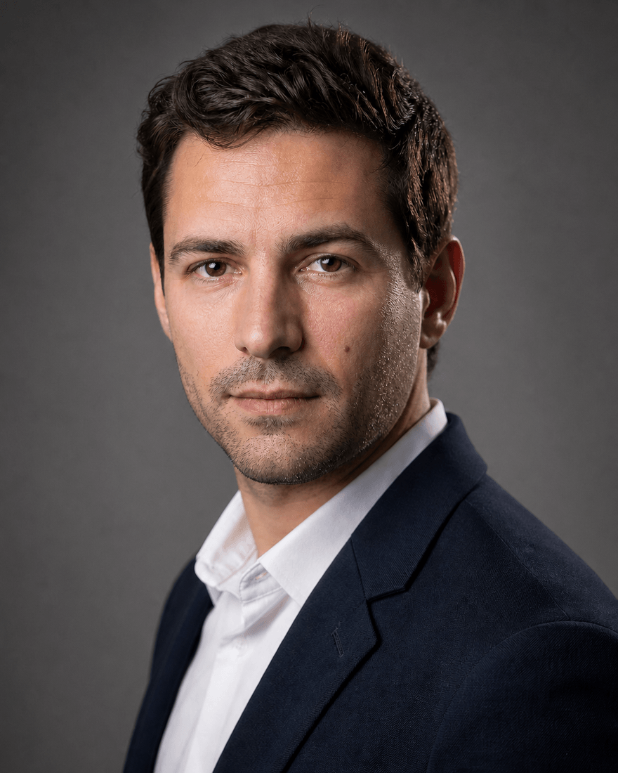 Derselbe Mann als Studioporträt vor mittelgrauem Hintergrund, weiches Licht von links,
            dunkelblaues Sakko über weißem Hemd, ruhiger Blick in die Kamera