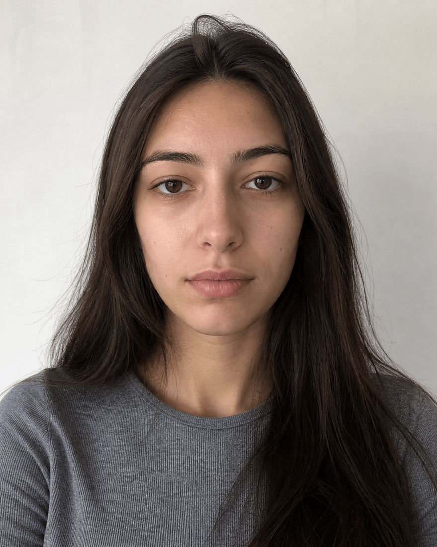 Ungeschminktes Portrait einer jungen Frau frontal vor einer weißen Wand im Tageslicht, neutraler Gesichtsausdruck
